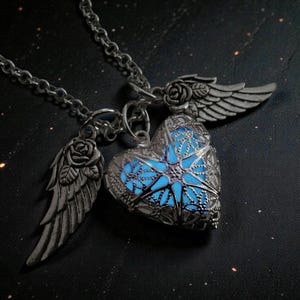 Pu&ograve; includere: Collana color argento con un ciondolo a forma di cuore con interno blu luminoso. Il ciondolo presenta un intricato design a filigrana ed &egrave; affiancato da due ali con dettagli di rose. Presentata su uno sfondo scuro.