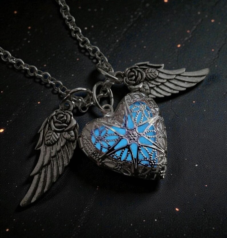 Pu&ograve; includere: Collana color argento con un ciondolo a forma di cuore con interno blu luminoso. Il ciondolo presenta un intricato design a filigrana ed &egrave; affiancato da due ali con dettagli di rose. Presentata su uno sfondo scuro.