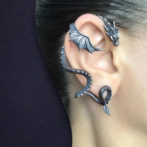 May include: A silver-toned ear cuff featuring a dragon design. The cuff wraps around the ear with a dragon head at the top, a wing detail, and a serpentine body that curves down the ear. The jewelry has intricate detailing.