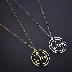 May include: Two pendant necklaces with a circular design. One necklace is gold-colored, the other is silver-colored. Each pendant features a stylized symbol within a circular frame, suspended from a delicate chain. The background is a dark, solid color.