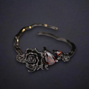 May include: A dark gray metal cuff bracelet featuring a rose and gemstone design. The bracelet has a large rose, a teardrop-shaped red gemstone, and a smaller red gemstone. The bracelet is open at the top.