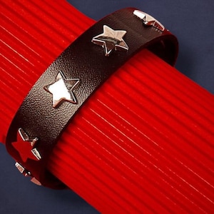 May include: A black leather bracelet with silver star studs. The bracelet is shown against a red background. The stars are evenly spaced around the bracelet.