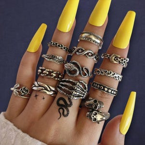 May include: A hand adorned with multiple silver rings featuring diverse designs such as a rose, a leaf, a snake, a skeleton hand, and skulls. The nails are painted bright yellow. A small snake tattoo is visible.