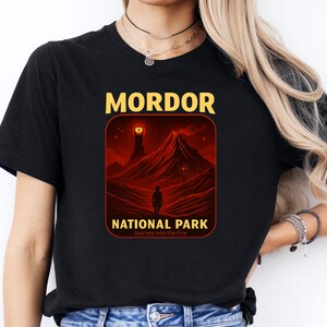 May include: Black t-shirt featuring a graphic of a red landscape with a silhouette of a person walking towards a mountain. The text "MORDOR NATIONAL PARK" is displayed in yellow and white, with "Journey Into the Fire" below.