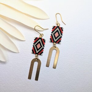 May include: Handmade earrings with a geometric pattern. The earrings have a rectangular beaded top in black, red, and white, with a gold-coloured U-shaped charm hanging below. The earrings have gold-coloured ear wires.