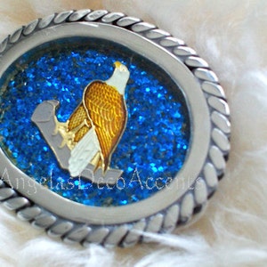 May include: Oval silver-toned belt buckle with a detailed eagle design. The eagle has gold and white feathers, perched on a gray branch against a blue glitter background. The buckle has a rope-like border and the text "Angelas Deco Accents".