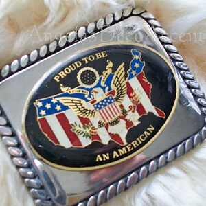 May include: Silver belt buckle with a black and gold oval design. The oval features a map of the United States with an eagle and the words "Proud to be an American".