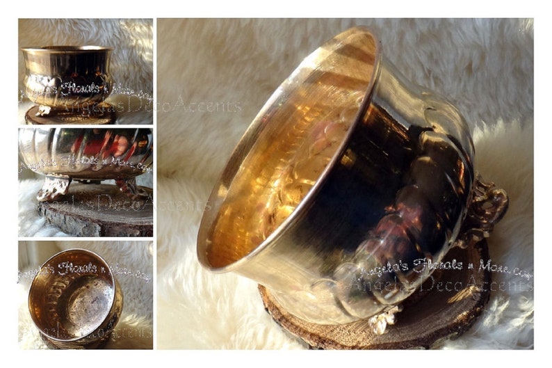 Op de afbeelding: Een metallic goudkleurige decoratieve kom met een brede opening, rustend op een houten schijf. De kom heeft een gedetailleerd, gestructureerd ontwerp en wordt ondersteund door sierlijke voetjes. De afbeelding bevat de tekst "Angela's Florals & More".