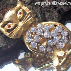 May include: A gold-toned cat brooch with red rhinestone eyes and a body encrusted with clear rhinestones. The brooch is set against a textured, dark brown background. The text "AngelasDecoAccents" is visible.