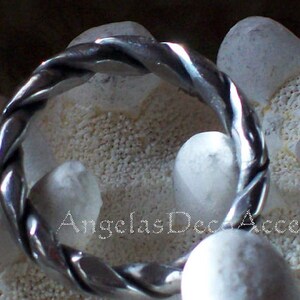 K&ouml;nnte beinhalten: Ein silberfarbener Ring mit einem gedrehten Seildesign. Der Ring ist vor einem Hintergrund aus wei&szlig;en Steinen und einer strukturierten Oberfl&auml;che platziert. Der Text "AngelasDecAccents" ist sichtbar.