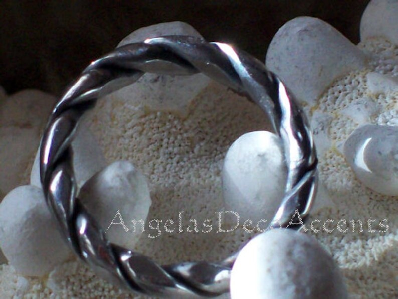 K&ouml;nnte beinhalten: Ein silberfarbener Ring mit einem gedrehten Seildesign. Der Ring ist vor einem Hintergrund aus wei&szlig;en Steinen und einer strukturierten Oberfl&auml;che platziert. Der Text "AngelasDecAccents" ist sichtbar.