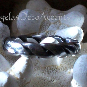 K&ouml;nnte beinhalten: Ein dunkles, geflochtenes Metallarmband mit poliertem Finish. Das Armband wird auf einem Bett aus wei&szlig;en, strukturierten Steinen pr&auml;sentiert. Der Text "Angelas Deco Accents" ist in der oberen linken Ecke sichtbar.
