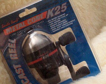 May include: A South Bend Metal Cone K25 cast reel in its original packaging. The reel is black and silver with red accents. The packaging includes text detailing features like all-metal gearing and a dual-grip power handle.