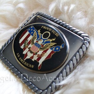May include: Silver belt buckle with a black background and a colourful image of the United States with an eagle and the words "Proud to be an American".