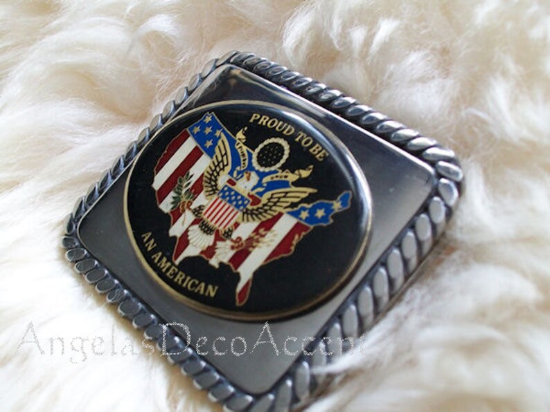 May include: Silver belt buckle with a black background and a colourful image of the United States with an eagle and the words "Proud to be an American".