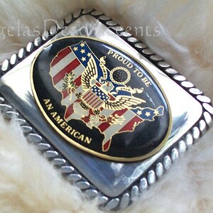May include: Silver belt buckle with a black and gold oval design. The oval features a stylised American flag with an eagle and the words "Proud to be an American".