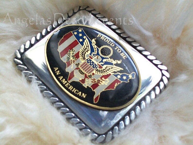 May include: Silver belt buckle with a black and gold oval design. The oval features a stylised American flag with an eagle and the words "Proud to be an American".