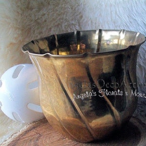 May include: A gold-colored, fluted vase with a wavy rim, next to a white plastic ball with cut-out holes. The vase has a reflective surface. The text "Angela's Deco Accents" and "Angela's Florals y More.com" is visible.