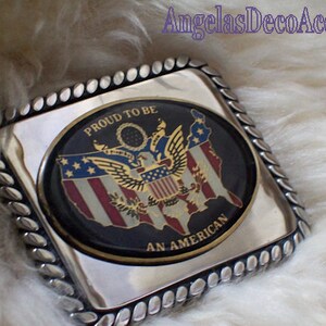 May include: A silver belt buckle with a black oval design featuring a gold eagle and the American flag. The text "Proud to be an American" is written around the oval.