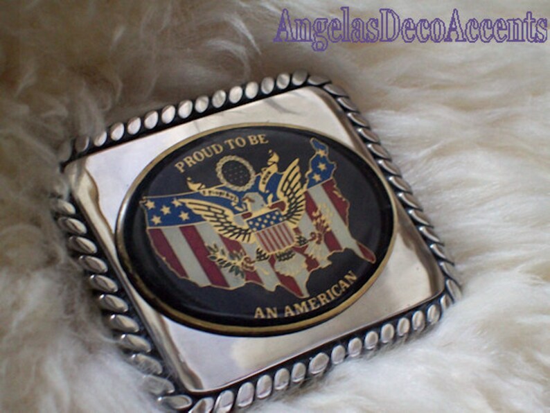 May include: A silver belt buckle with a black oval design featuring a gold eagle and the American flag. The text "Proud to be an American" is written around the oval.