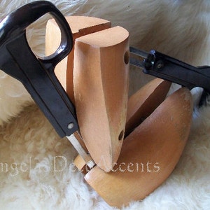 K&ouml;nnte beinhalten: Schuhspanner aus Holz mit schwarzen Griffen und Metallkomponenten. Die Schuhspanner sind hellbraun und sollen die Form der Schuhe erhalten. Das Bild ist vor einem wei&szlig;en, flauschigen Hintergrund aufgenommen.