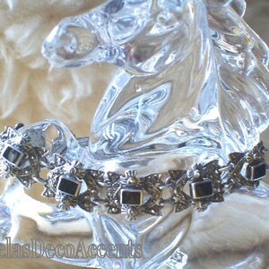 May include: A silver-toned bracelet with black rectangular stones is draped over a clear glass horse figurine. The bracelet has an ornate design with small decorative elements. The background is a soft, neutral color.