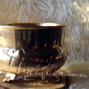 Op de afbeelding: Een metallic goudkleurige plantenbak met een brede, afgeronde komvorm. De plantenbak staat op een houten voet en heeft decoratieve details rond de rand. De achtergrond is een zacht, getextureerd, gebroken wit oppervlak.