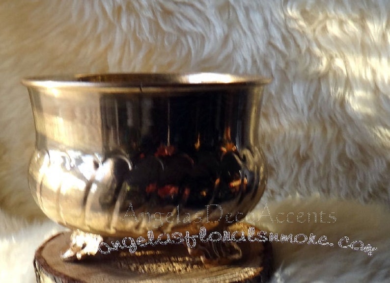 Op de afbeelding: Een metallic goudkleurige plantenbak met een brede, afgeronde komvorm. De plantenbak staat op een houten voet en heeft decoratieve details rond de rand. De achtergrond is een zacht, getextureerd, gebroken wit oppervlak.