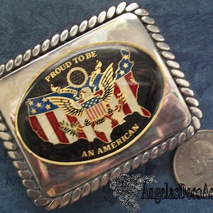 May include: A silver belt buckle with a rope-like border. The buckle features a black oval with a gold border. Inside the oval is a colourful image of the United States with an eagle and the words "Proud to be an American".