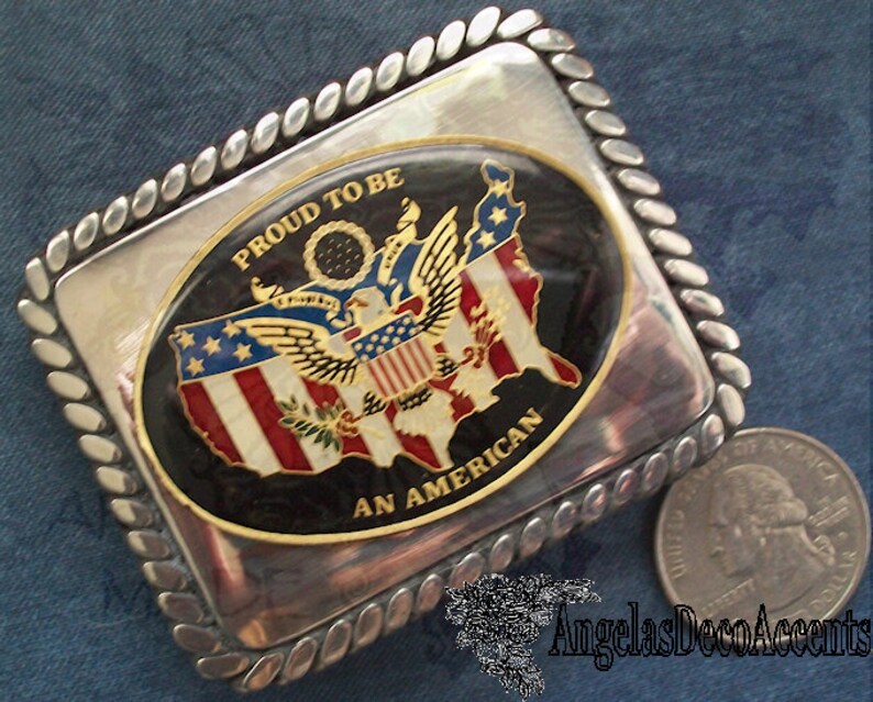 May include: A silver belt buckle with a rope-like border. The buckle features a black oval with a gold border. Inside the oval is a colourful image of the United States with an eagle and the words "Proud to be an American".