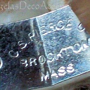 K&ouml;nnte beinhalten: Nahaufnahme eines gl&auml;nzenden, silbernen Metallobjekts mit eingraviertem Text. Der Text enth&auml;lt "U.S. PAT." und "BROCKTON MASS". Das Objekt hat eine gebogene Form mit einem Loch auf einer Seite. Das Bild ist leicht unscharf.