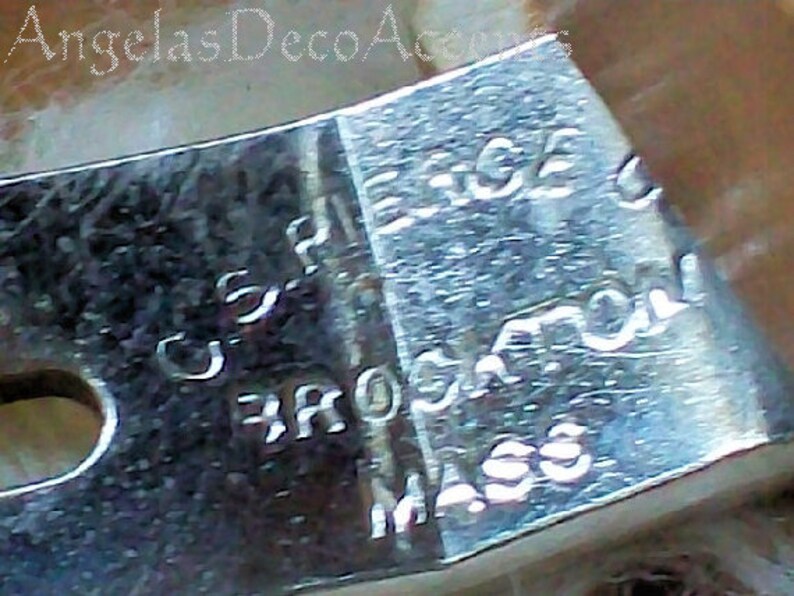 K&ouml;nnte beinhalten: Nahaufnahme eines gl&auml;nzenden, silbernen Metallobjekts mit eingraviertem Text. Der Text enth&auml;lt "U.S. PAT." und "BROCKTON MASS". Das Objekt hat eine gebogene Form mit einem Loch auf einer Seite. Das Bild ist leicht unscharf.
