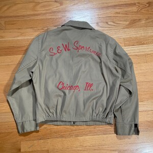 May include: A vintage green work jacket with red embroidered text on the back that reads "S&W Sportsmen Chicago, Ill."