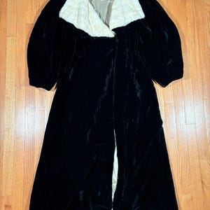May include: A long, black velvet coat with a cream-colored, textured collar. The coat has a flowing design and a split front. The collar has a label with text. The coat is laid out on a wooden surface.