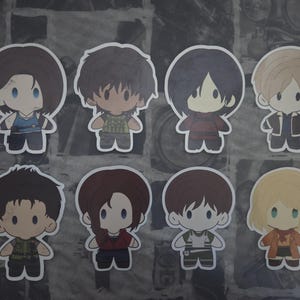 May include: A set of eight chibi-style character stickers. Each sticker depicts a different character with large eyes and simplified features. The characters are dressed in various outfits, with a variety of hair colours and styles.