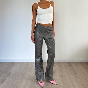 Puede incluir: Pantalones grises de imitación de piel de serpiente con pernera acampanada. Los pantalones se combinan con una camiseta sin mangas blanca con tirantes finos y tacones rosas de punta. Los pantalones tienen cierre de botón y cremallera.