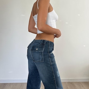 May include: A white spaghetti strap tank top paired with dark wash denim cargo pants. The jeans feature large pockets on the back and a button closure. The pants have a slight flare at the bottom.