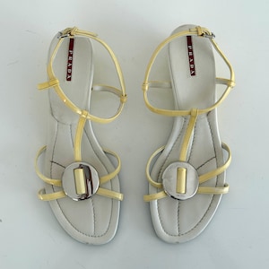 May include: A pair of pale yellow patent leather sandals with thin ankle straps and a T-strap design. Each sandal features a large, circular silver-tone metal ornament with a central yellow bar. The white insoles are branded with the Prada logo.