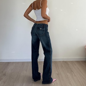 May include: Dark wash denim jeans with a wide leg and two cargo pockets on the back. The jeans have a low waist and are paired with a white tank top. The model is wearing pink pointed-toe heels.