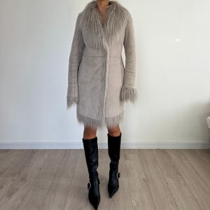 May include: A light beige coat with a faux fur collar and trim. The coat has long sleeves and a knee-length hem. The person is wearing black knee-high boots with pointed toes and silver buckles. The coat is a stylish outerwear garment.