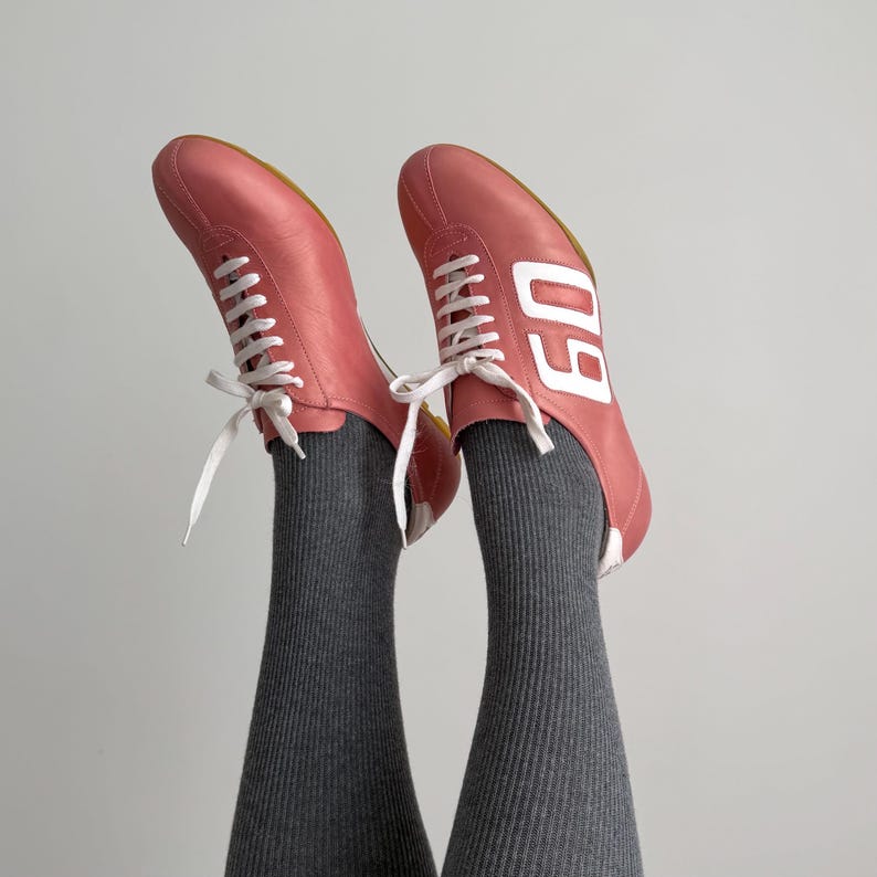 May include: A pair of pink leather sneakers with white laces and the number "60" printed in white on the side. The shoes are worn with gray ribbed socks. The background is a solid light gray.