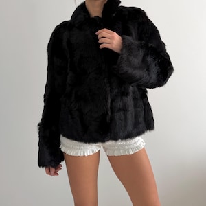 May include: A black fur jacket with a collared neckline and long sleeves. The jacket is worn over white shorts. The fur appears soft and plush, creating a luxurious look. The jacket is buttoned up.
