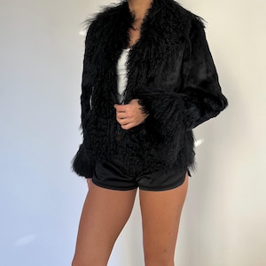 Peut inclure: Veste en fausse fourrure noire &agrave; l'aspect textur&eacute; et hirsute. La veste a un large col et des manches longues. Le mod&egrave;le porte un short noir et un haut blanc. Le fond est un mur blanc uni.
