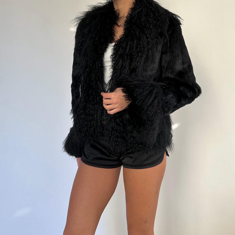 Peut inclure: Veste en fausse fourrure noire &agrave; l'aspect textur&eacute; et hirsute. La veste a un large col et des manches longues. Le mod&egrave;le porte un short noir et un haut blanc. Le fond est un mur blanc uni.