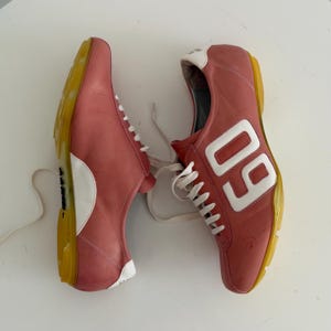 May include: Pair of pink sneakers with white laces and accents. The sneakers have a yellow sole and the number "09" in white on the side. The shoes are made of leather.