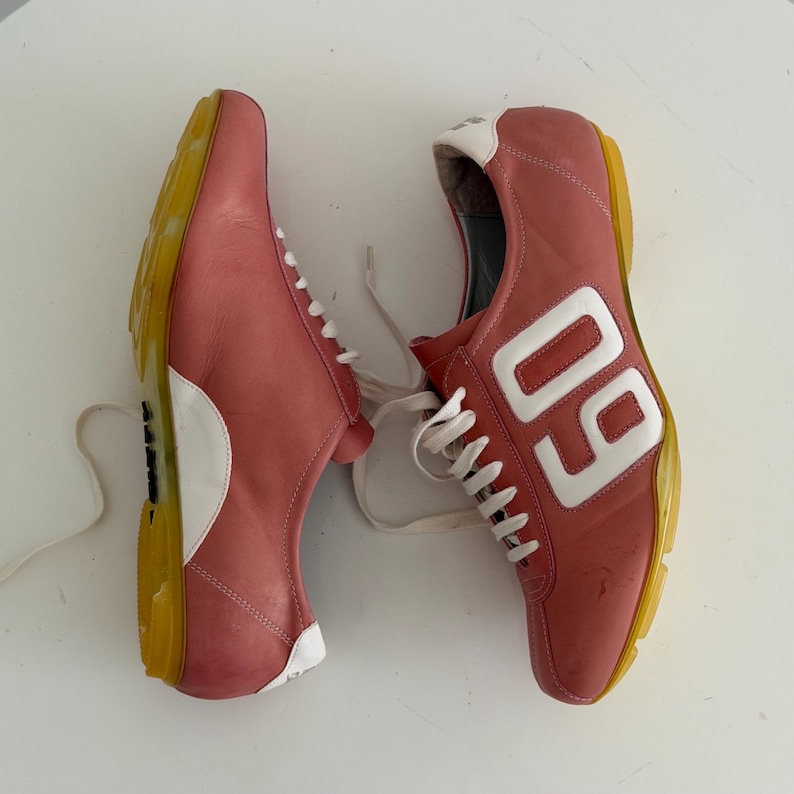 May include: Pair of pink sneakers with white laces and accents. The sneakers have a yellow sole and the number "09" in white on the side. The shoes are made of leather.