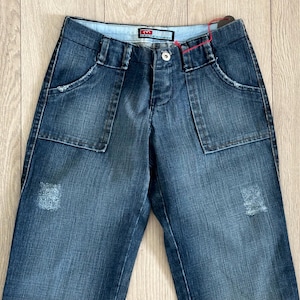 May include: Blue denim jeans with large front pockets and a button closure. The jeans have a distressed patch on the front and a small logo tag near the waistband. The jeans are laid flat on a light wood surface.