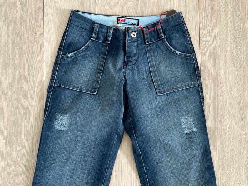 May include: Blue denim jeans with large front pockets and a button closure. The jeans have a distressed patch on the front and a small logo tag near the waistband. The jeans are laid flat on a light wood surface.