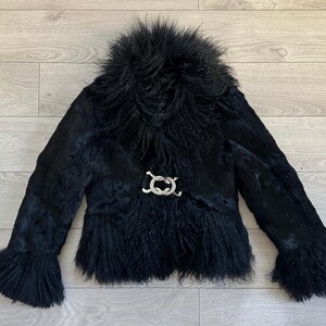 Peut inclure: Veste en fourrure noire avec un col duveteux et des manches &eacute;vas&eacute;es. La veste est orn&eacute;e d'un fermoir d&eacute;coratif argent&eacute; &agrave; la taille. Le v&ecirc;tement est fait d'une mati&egrave;re textur&eacute;e et est pos&eacute; sur une surface en bois.