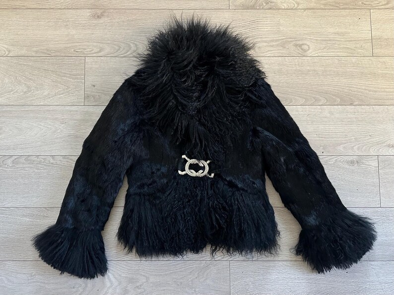 Peut inclure: Veste en fourrure noire avec un col duveteux et des manches &eacute;vas&eacute;es. La veste est orn&eacute;e d'un fermoir d&eacute;coratif argent&eacute; &agrave; la taille. Le v&ecirc;tement est fait d'une mati&egrave;re textur&eacute;e et est pos&eacute; sur une surface en bois.